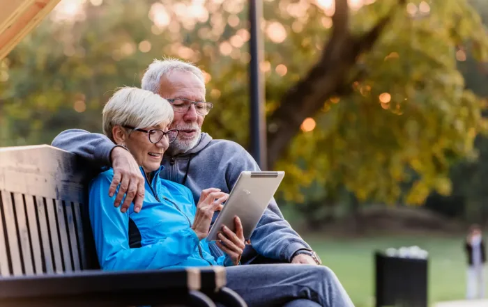 Smiling retired couple sitting on a park bench using a tablet to review their retirement finances